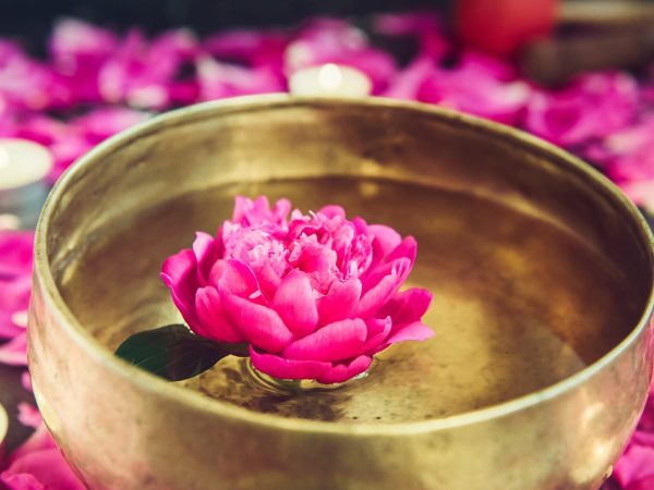 Tibetan singing bowl with floating in water pink peony flower. Burning candles and petals on the black stone background. Meditation and Relax. Exotic massage. Vertical. Selective focus. Copy space